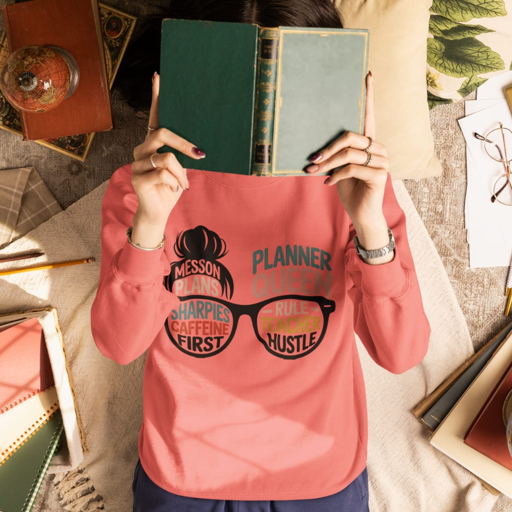 Person wearing a pink sweatshirt with text, holding a book over their face in a desk setting.