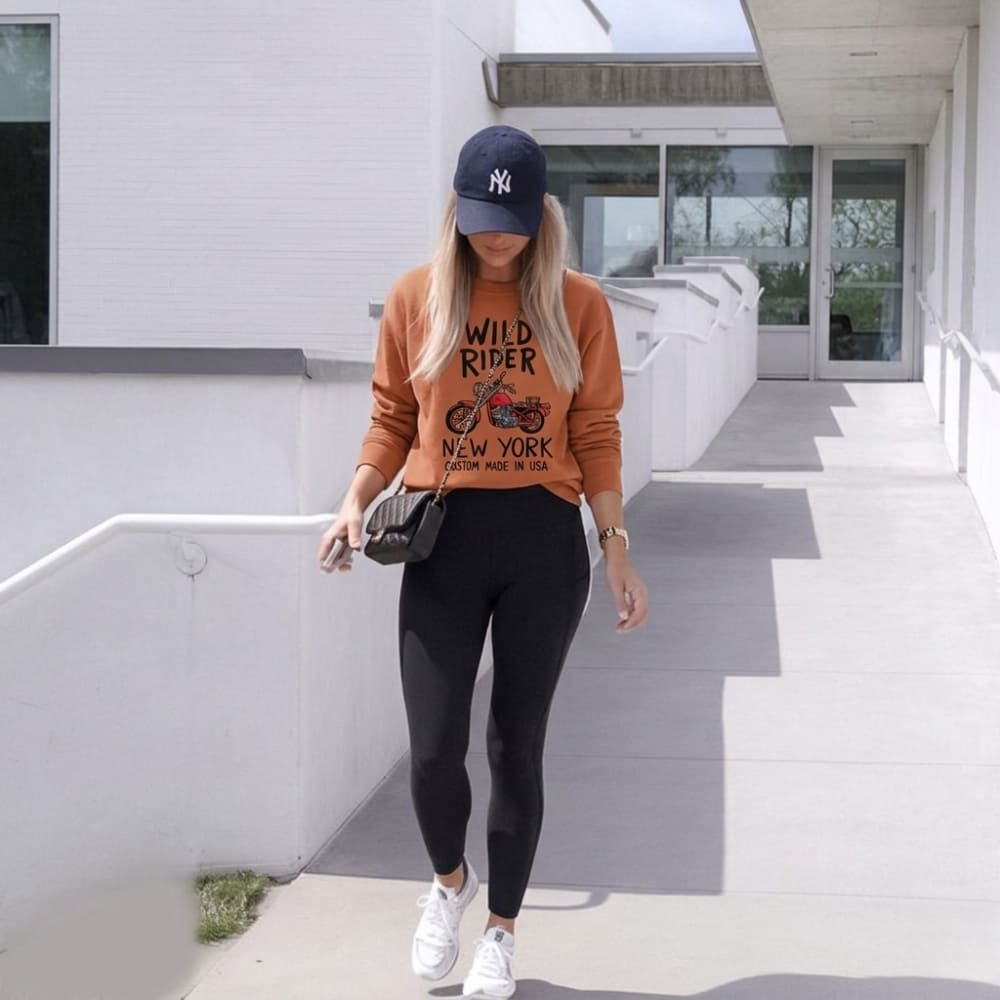 Woman wearing an orange sweatshirt with text and graphics, black leggings, white sneakers, and a blue cap in an outdoor setting.