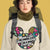 Person wearing a beige sweatshirt with colorful graphic and text, standing against a yellow background.