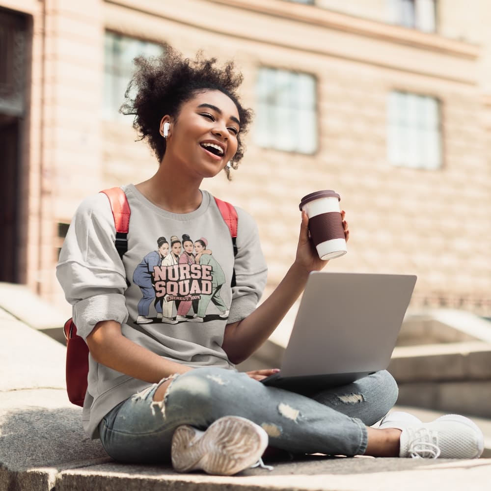 Woman sitting outdoors with a laptop and coffee, wearing a 'Nurse Squad' t-shirt.