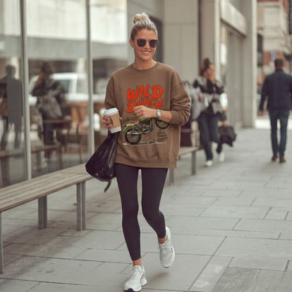 Woman walking on a city street wearing a brown sweatshirt with 'Wild Bunch' text, dark leggings, and sunglasses.