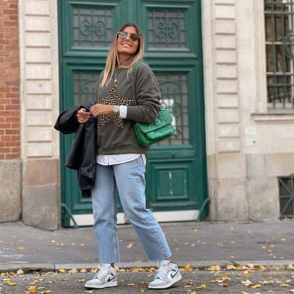 Woman standing in front of a green door wearing a gray sweater, leopard print top, and blue jeans.