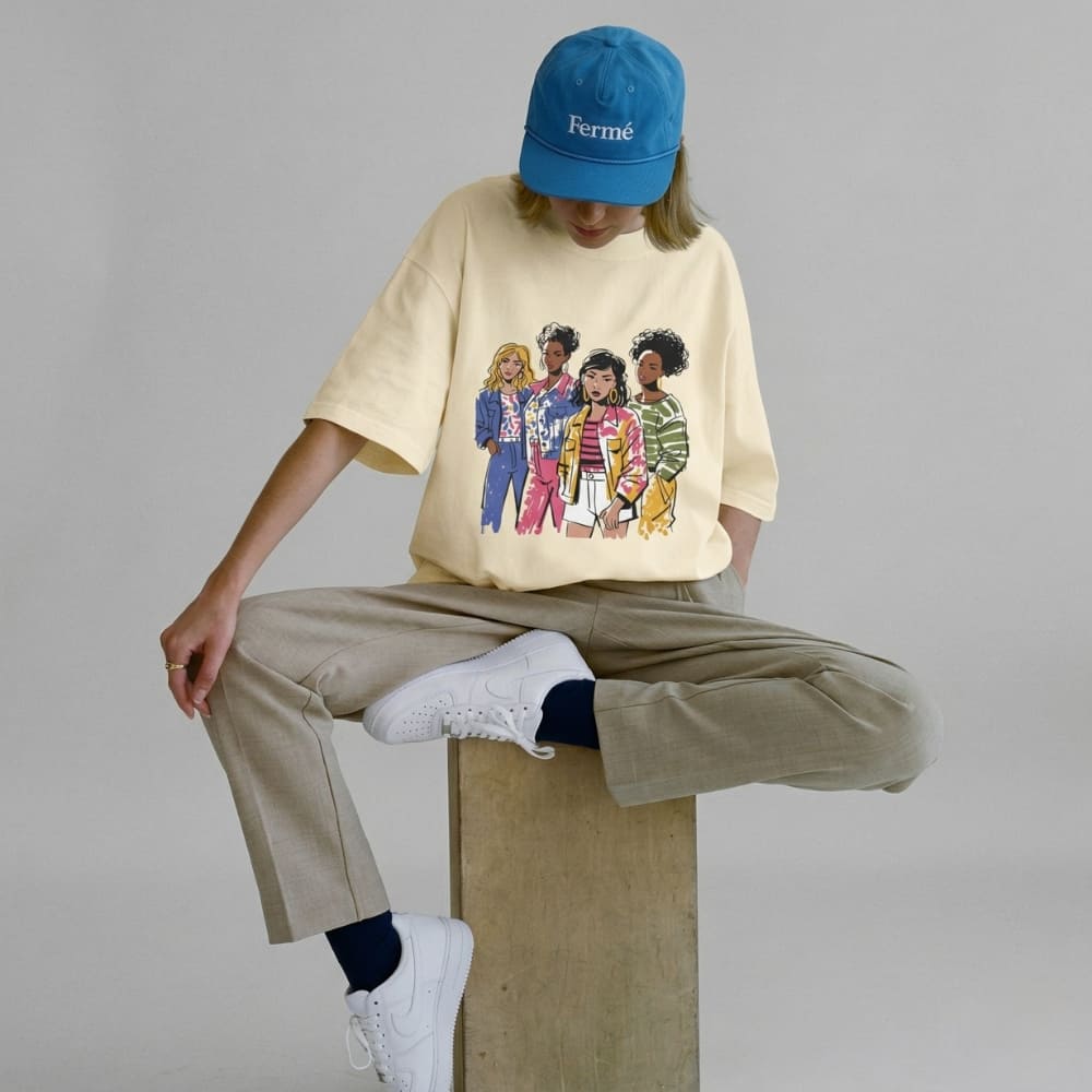 Person wearing a beige t-shirt with a graphic design, beige pants, white sneakers, and a blue cap sitting on a block.