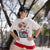 Woman wearing a white t-shirt with a graphic design and text, standing outdoors with trees in the background.