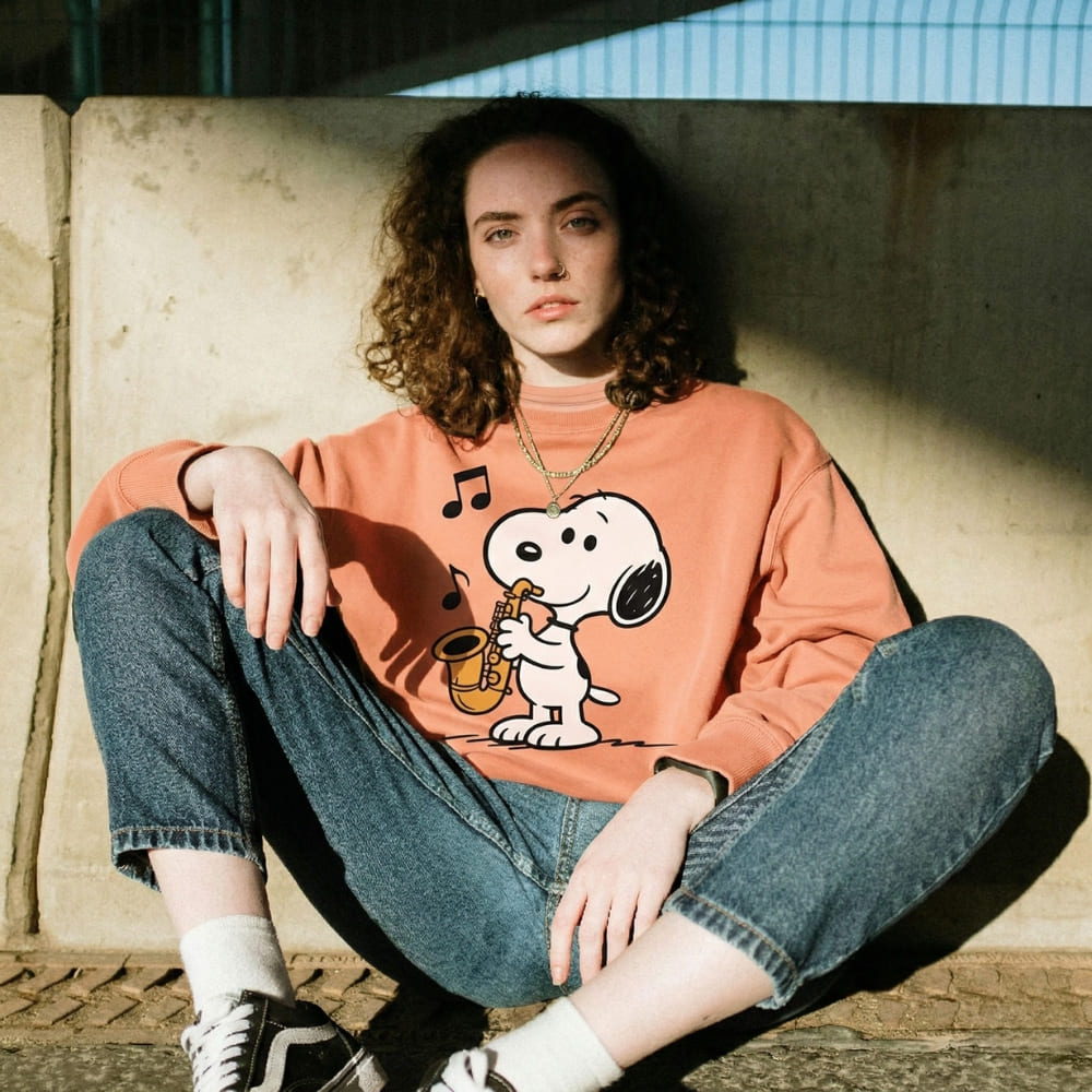 Person wearing a Snoopy peach-colored sweatshirt sitting on a concrete surface.