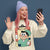 Person wearing a beige sweatshirt with a cartoon character design, holding a phone, and waving against a dark blue background.