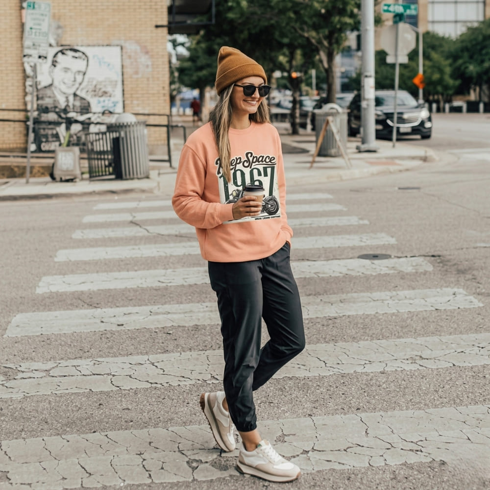 Person wearing a peach-colored sweatshirt with text, black pants, and a beanie, crossing a street.