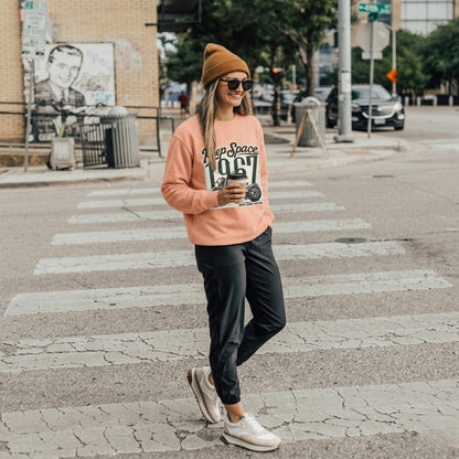 Person wearing a peach-colored sweatshirt with text, black pants, and a beanie, crossing a street.