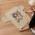 Beige t-shirt with a graphic design of a woman holding a coffee cup on a wooden floor.