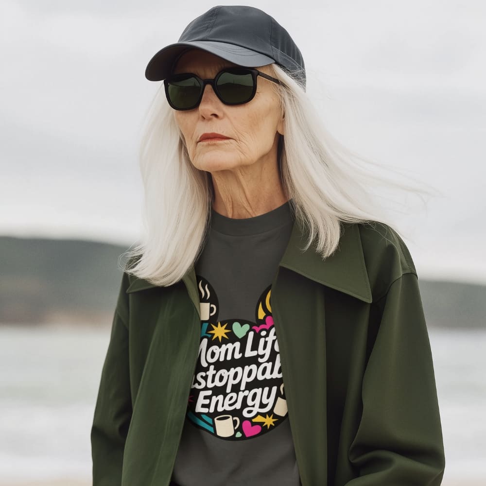 Woman wearing sunglasses, cap, and jacket with a graphic t-shirt by a body of water.