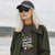 Woman wearing sunglasses, cap, and jacket with a graphic t-shirt by a body of water.