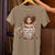 T-shirt with a graphic of a person in a coffee cup and text 'Don't ask me me anything before 2 cup' on a hanger.