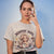 Woman wearing a t-shirt with a graphic and text 'Mommin' Hard. Caffeinating Harder.' against a blue background