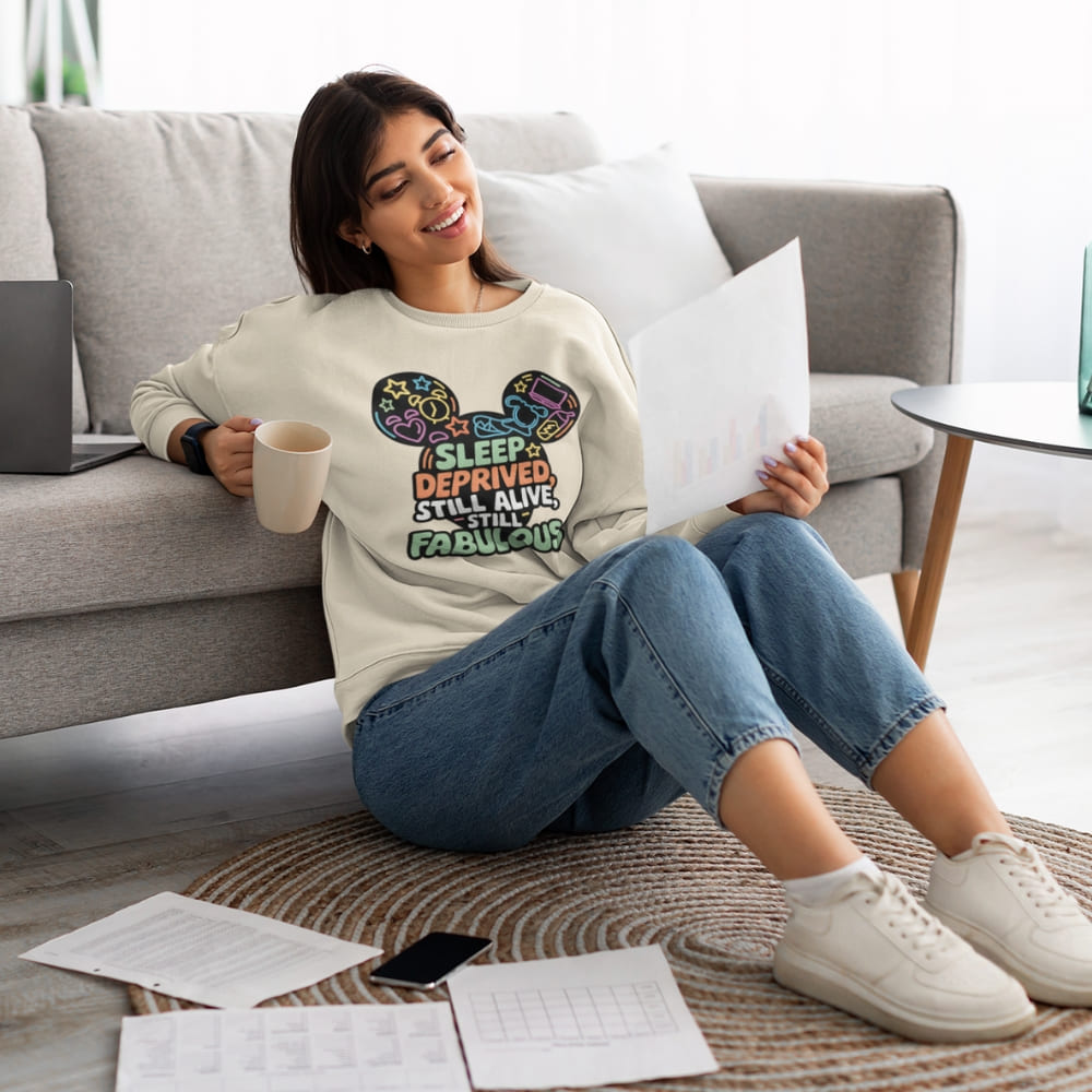 Woman sitting on the floor with a coffee cup, holding a paper, wearing a sweatshirt with colorful text design.