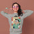 Woman wearing a sweatshirt with a cartoon character design, holding a skateboard against a brown background