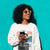 Woman wearing a white sweatshirt with a graphic design, holding a phone against a teal background