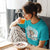 Woman in a blue t-shirt with a graphic design, sitting at a table with a bowl of cereal and a glass of orange juice.