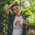 Person wearing a t-shirt with a graphic design and text, standing in a forest.