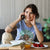 Woman sitting at a table with food and a book, wearing a blue t-shirt with a graphic design.