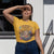Woman wearing a yellow t-shirt with a graphic design and text, standing outdoors.