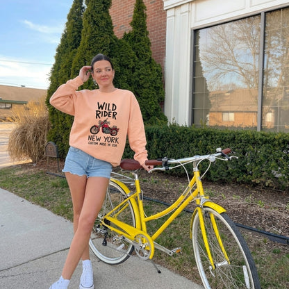 Woman in peach-colored sweatshirt and denim shorts standing next to a yellow bicycle outdoors.
