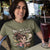Woman wearing a t-shirt with a coffee-themed graphic and text, sitting at a table with a drink.