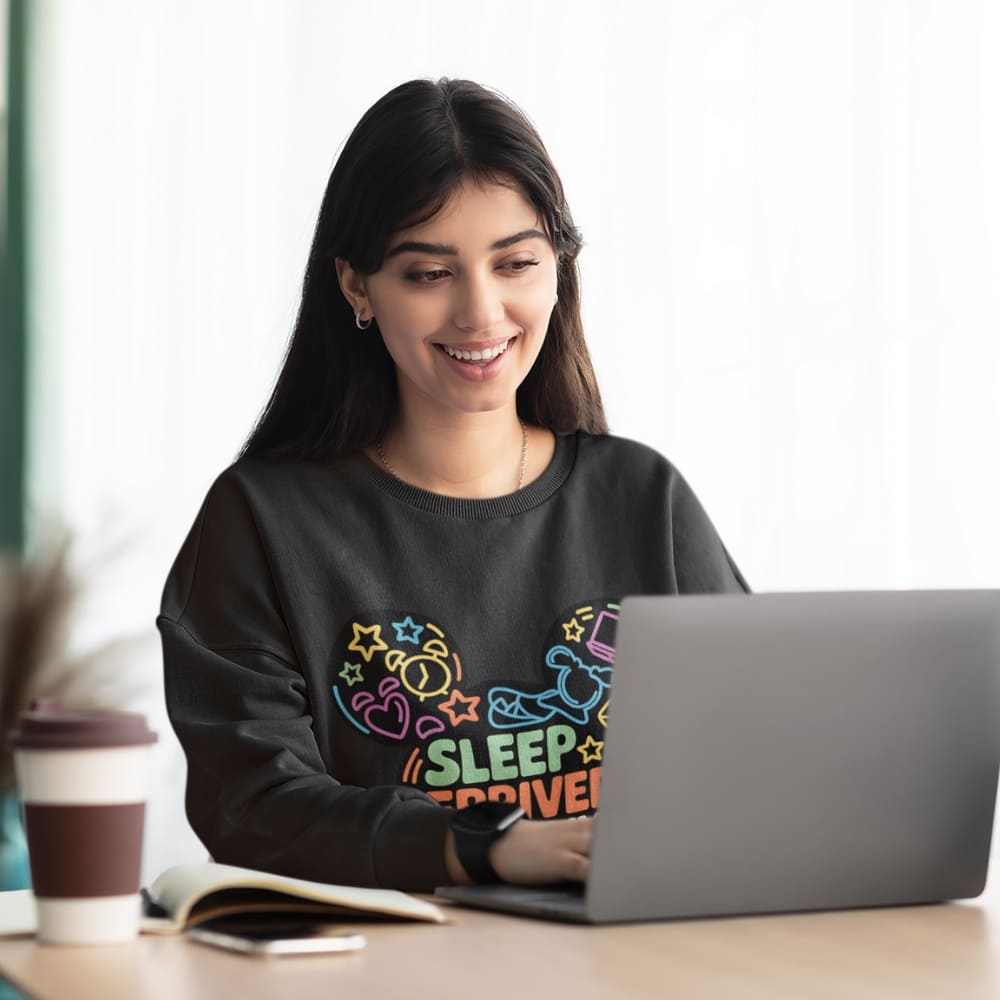 Woman wearing a black sweatshirt with colorful text, sitting at a desk with a laptop.