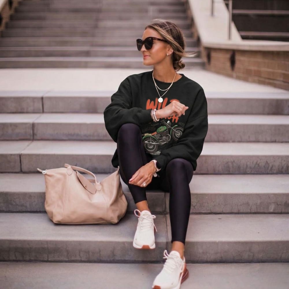 Woman sitting on steps wearing a black sweatshirt, leggings, and white sneakers with a beige bag beside her.