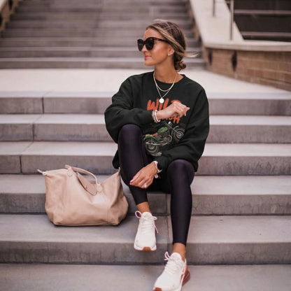 Woman sitting on steps wearing a black sweatshirt, leggings, and white sneakers with a beige bag beside her.