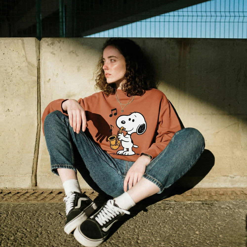 Person wearing a orange sweatshirt with a Snoopy graphic sitting against a wall.
