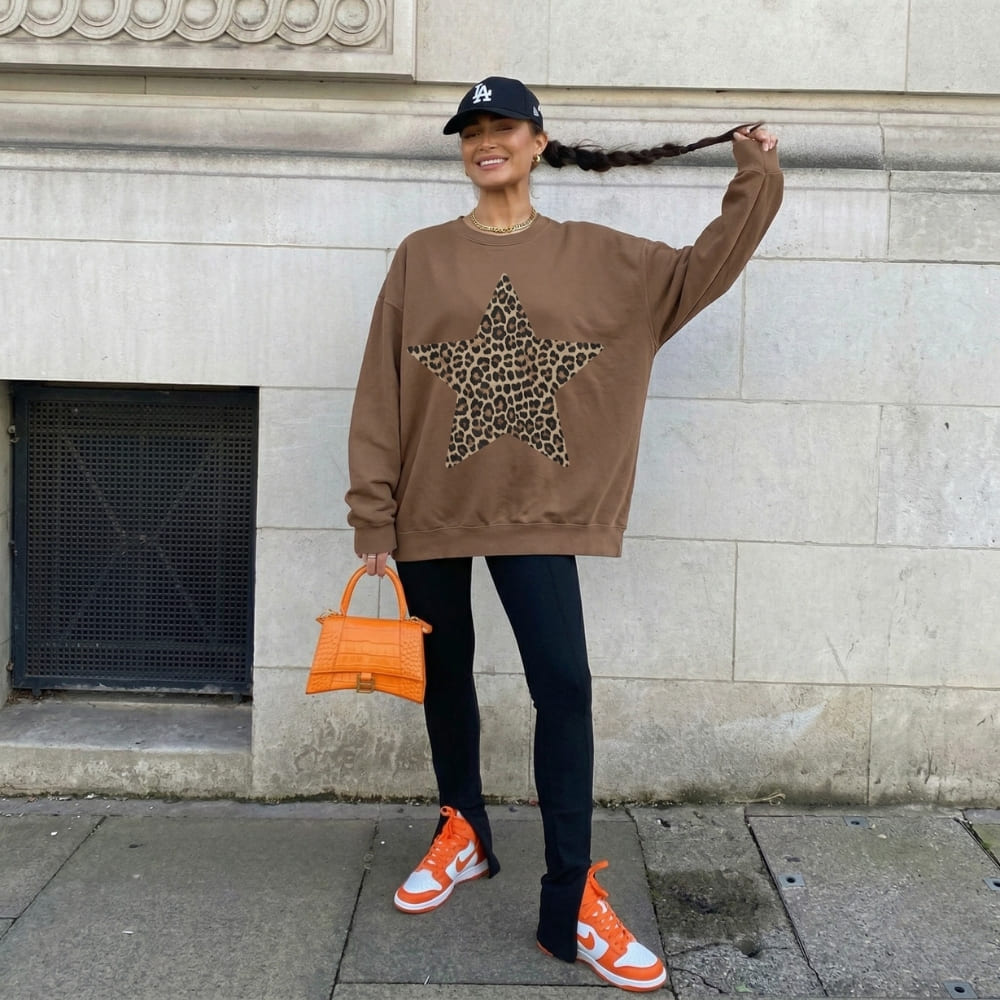 Woman wearing a brown sweatshirt with a leopard print star, black leggings, orange sneakers, and an orange handbag, standing against a light-colored wall.