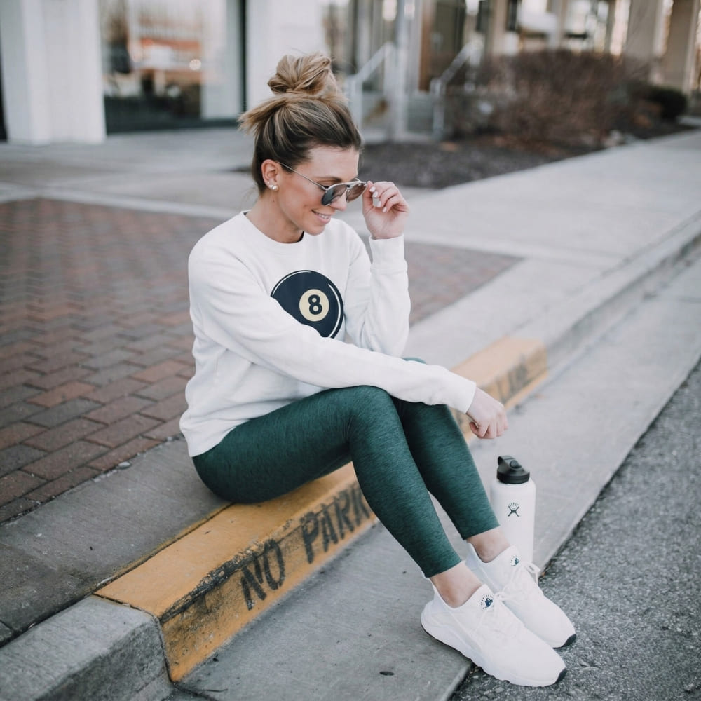 Woman sitting on a curb wearing a white sweatshirt with a logo, green leggings, and white sneakers.