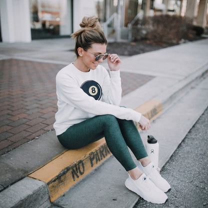 Woman sitting on a curb wearing a white sweatshirt with a logo, green leggings, and white sneakers.