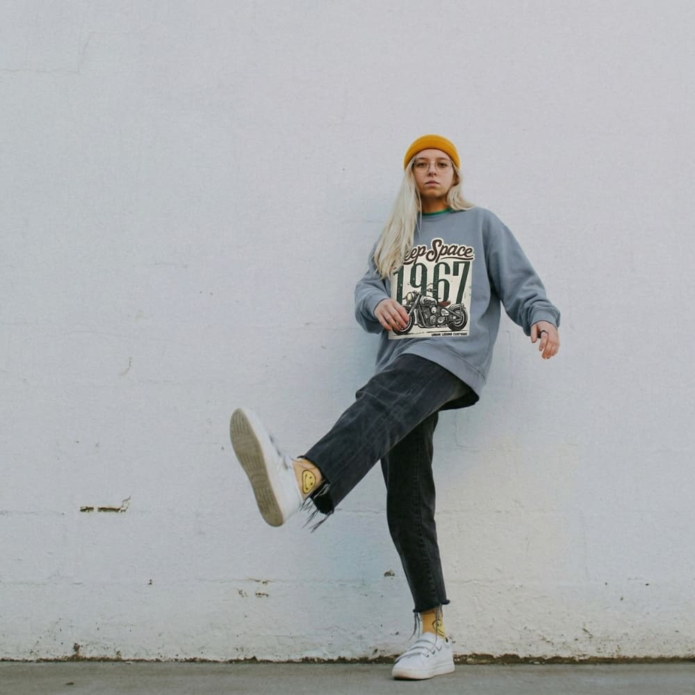 Person wearing a blue jean sweatshirt with text, black pants, and white sneakers against a white wall.