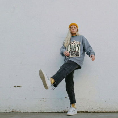 Person wearing a blue jean sweatshirt with text, black pants, and white sneakers against a white wall.