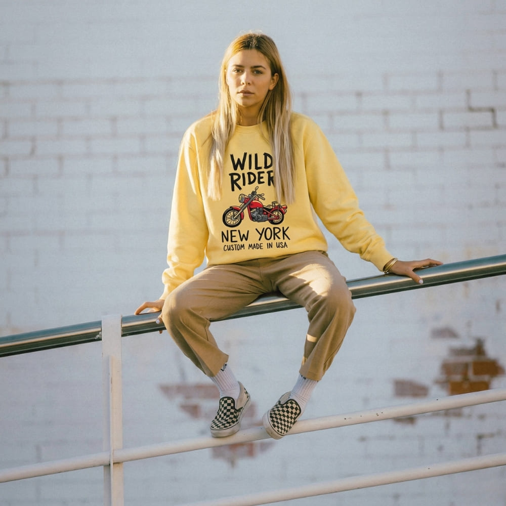 Person wearing a yellow sweatshirt with 'Wild Rider New York' text, sitting on a railing.