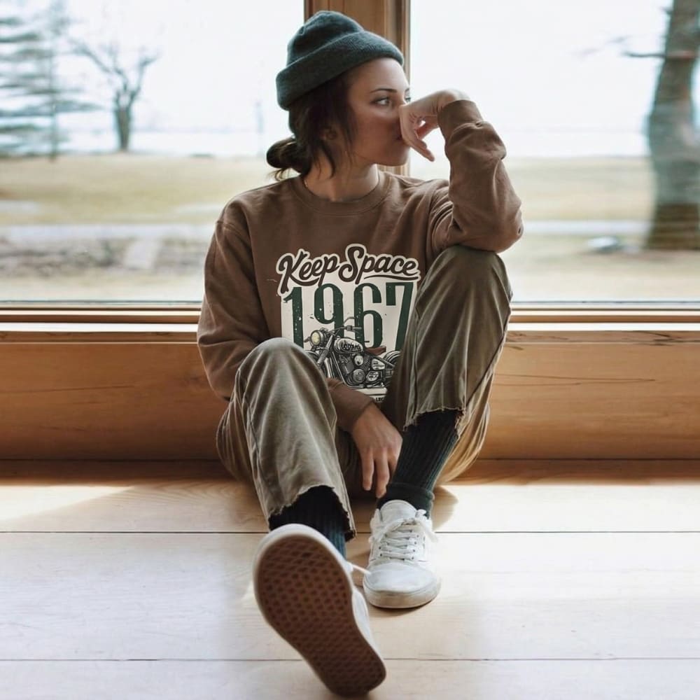 Person wearing a brown sweatshirt with text, sitting by a window.