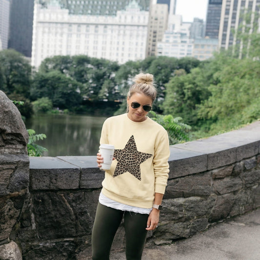Woman wearing a beige sweater with a leopard print star, holding a coffee cup, standing in an urban park setting.