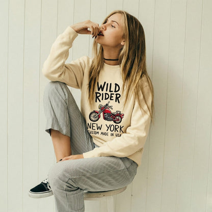 Woman wearing a ivory 'Wild Rider New York' sweatshirt sitting against a light wooden paneled wall.