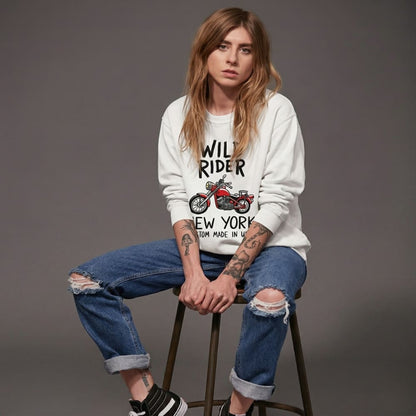 Woman sitting on a stool wearing a white sweatshirt with text and graphics, blue jeans, and black shoes.