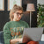 Woman wearing a green sweatshirt with text, using a laptop in a cozy room.