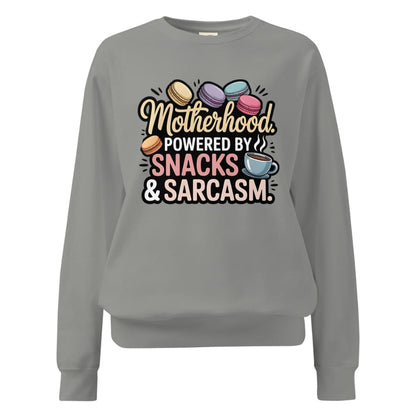 Sweatshirt design with text “Motherhood. Powered by Snacks & Sarcasm.” featuring colorful macarons and a steaming coffee cup on vintage-washed cotton fleece.