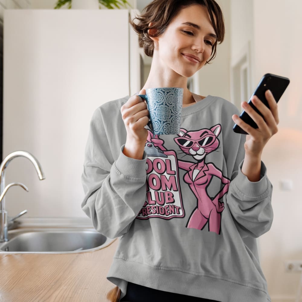 Woman in a kitchen wearing a sweatshirt with a pink Panther graphic, holding a mug and a phone.