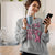 Woman in a kitchen wearing a sweatshirt with a pink Panther graphic, holding a mug and a phone.