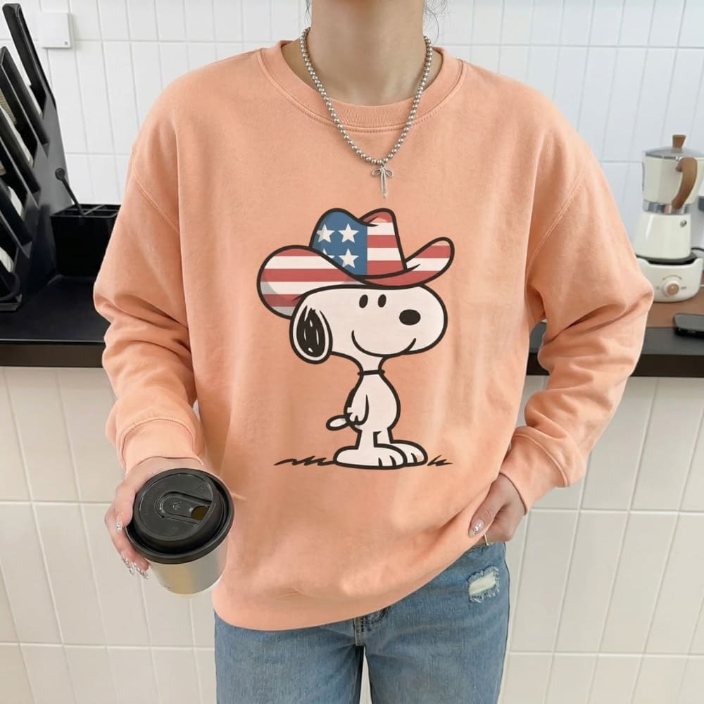 Person wearing a peach sweatshirt with a cartoon character holding a coffee cup in a kitchen.