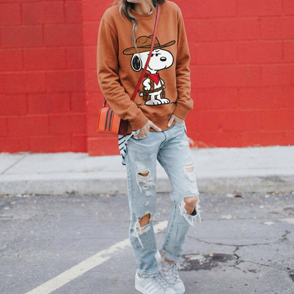 Person wearing a orange sweatshirt with a graphic design, light blue ripped jeans, and white sneakers against a red brick wall.