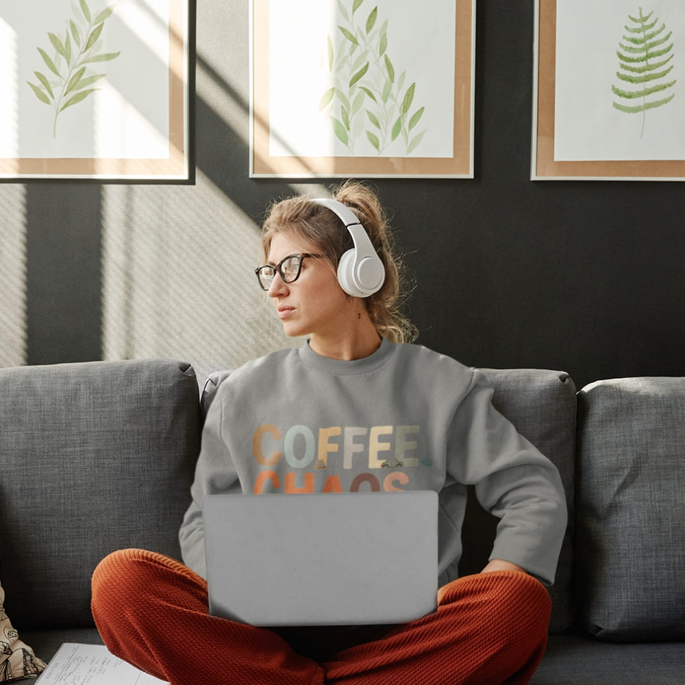 Person sitting on a couch with a laptop wearing a 'Coffee Fueled' sweatshirt, surrounded by framed plant art.