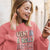 Woman wearing a pink sweatshirt with text, listening to music on earphones, and looking at her phone.