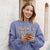 Woman wearing a purple sweatshirt with text, listening to music and looking at her phone.
