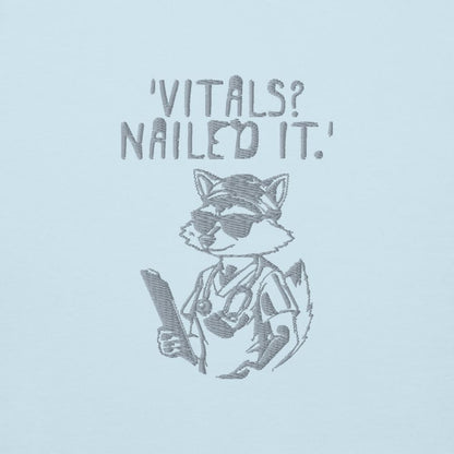 Raccoon nurse graphic wearing scrubs and sunglasses holding a clipboard under the quote “Vitals? Nailed It.” – printed on a black Cotton Heritage hoodie